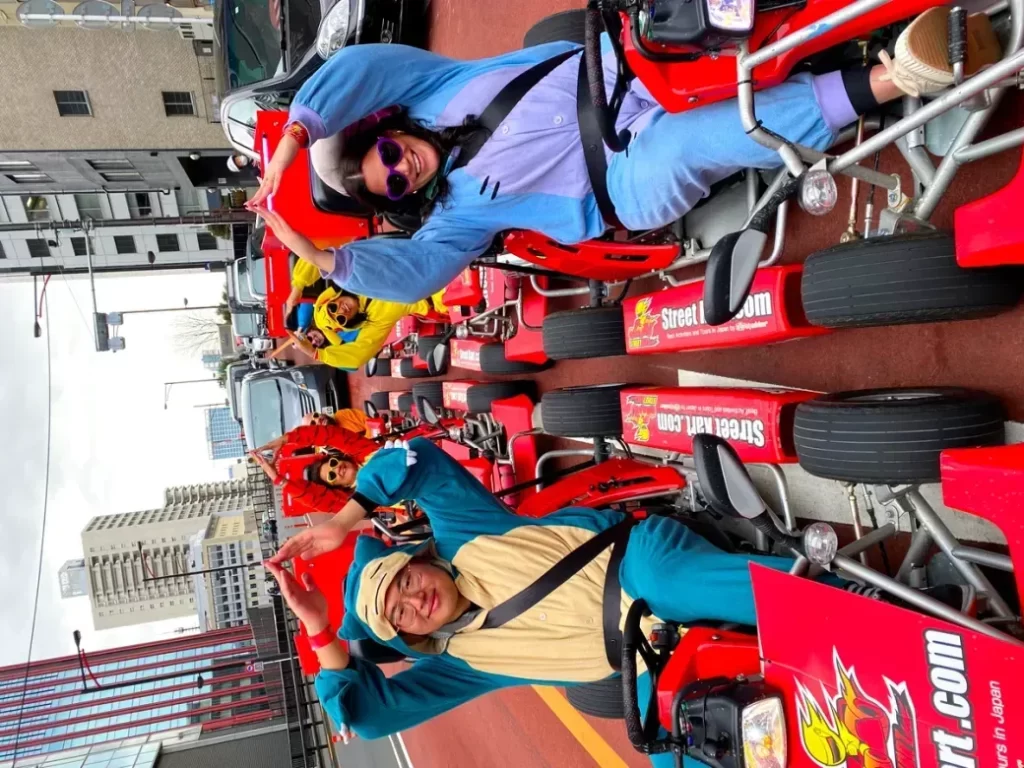 IMG_2936 - StreetKart Group of people in red go-karts on a city street, smiling and waving at the camera during a street event.”,
