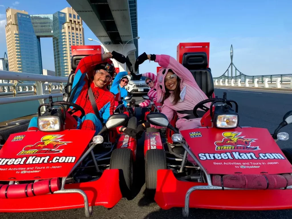 IMG_4112 - StreetKart Four people in red go-karts dressed as Spider-Man and friends making a heart shape with their arms on a bridge, city buildings in the background.